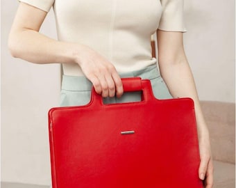 May include: A bright red, rectangular handbag with a top handle. The bag has a silver-colored metal accent near the top. The bag is held by a person wearing a cream-colored off-the-shoulder top and light blue pants.