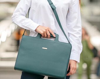 May include: Teal leather briefcase with a top handle and adjustable shoulder strap. The bag has a rectangular shape with a small gold-colored accent on the front. The person is wearing a white button-down shirt.