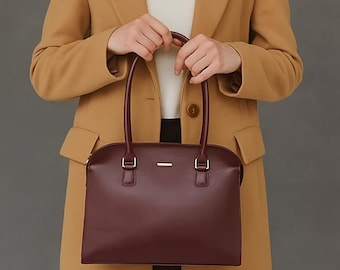 May include: A woman wearing a tan coat and holding a burgundy handbag with a rectangular shape and two handles. The bag has a silver bar detail and is made of a smooth material. The woman is wearing a white top.