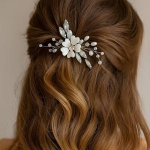 Peut inclure: Une barrette à cheveux délicate avec un motif floral blanc, un cristal central et des accents de cristaux transparents et vert pâle. La barrette est posée sur une base argentée et est placée dans des cheveux bruns.