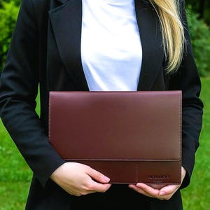 May include: A burgundy leather portfolio with a contrasting darker brown panel, held against a background of green grass. The portfolio has a stitched border and the text "M. MANAGER" is visible. The person is wearing a black blazer and a white shirt.