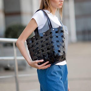 May include: A black woven leather tote bag with a rectangular shape. The bag has a woven pattern and is held by a person wearing a white t-shirt and blue jeans. The bag has a shoulder strap.