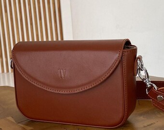 Può includere: Una borsa a tracolla in pelle marrone con chiusura a patta. La borsa ha una forma arrotondata e un design cucito. Una tracolla color argento è attaccata alla borsa. La lettera "W" è impressa sul davanti.