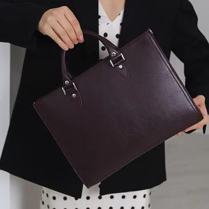 May include: A dark burgundy leather briefcase with a top handle and silver-tone hardware. The rectangular bag has a structured design and is held by a person wearing a black blazer and a white polka dot dress.