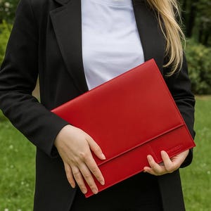 Op de afbeelding: Een felrode leren portfolio, rechthoekig met een klepsluiting, gehouden tegen een achtergrond van groen gras. De persoon draagt een zwarte blazer en een wit t-shirt.
