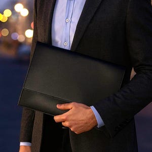 Op de afbeelding: Een zwarte leren portfolio, vastgehouden door een persoon die een donkergrijze blazer en een lichtblauw overhemd draagt. De portfolio heeft een eenvoudig, professioneel ontwerp, geschikt voor zakelijke of formele gelegenheden.