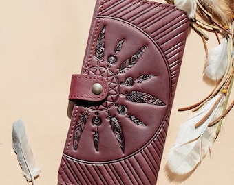May include: A burgundy leather wallet with a flap closure and a button clasp. The wallet features an embossed dreamcatcher design with feathers and geometric patterns. The background includes feathers and leather straps.