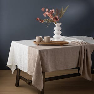 May include: A rectangular wooden table with a two-tone linen tablecloth. The tablecloth is white and tan. A wooden tray holds two beige mugs. A white vase with pink flowers sits on the table.