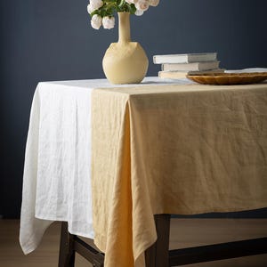 May include: A table setting with a white and tan linen tablecloth. A yellow vase holds white flowers. A stack of books and a decorative wooden plate are also on the table. The table is set against a dark blue background.