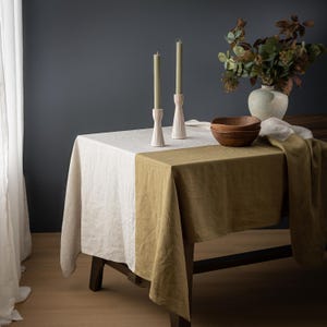 May include: A dining table set with a white and olive green linen tablecloth. Two light pink candle holders with green candles are on the table. A vase with greenery and two wooden bowls complete the setting.