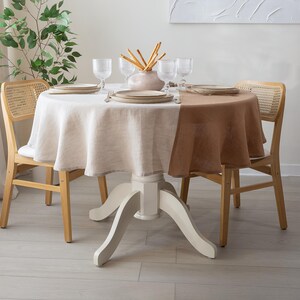 May include: A round dining table set for a meal. The table has a two-tone linen tablecloth, with a white and brown section. The table is set with plates, glasses, and a vase with breadsticks. Two wooden chairs with woven backs are on either side.