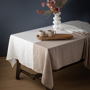 May include: A two-tone linen tablecloth in white and beige covers a wooden table. Two beige mugs sit on a wooden tray, next to a white vase with pink flowers. The table is set against a dark blue wall.