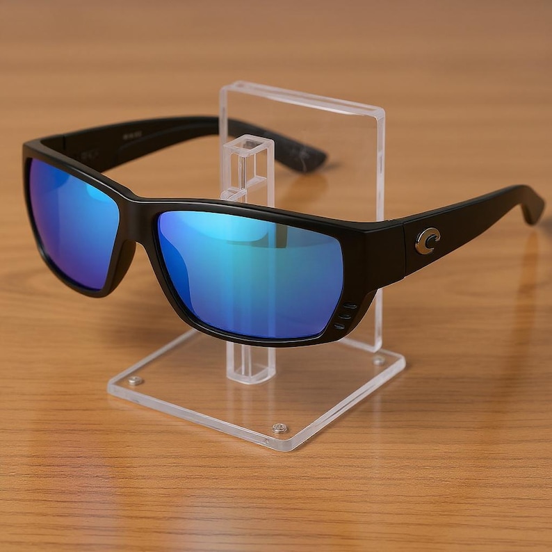 Puede incluir: Gafas de sol negras con lentes de espejo azules se exhiben en un soporte de acr&iacute;lico transparente. Las gafas de sol tienen una montura rectangular y un dise&ntilde;o curvo. Las lentes reflejan un tono azul.