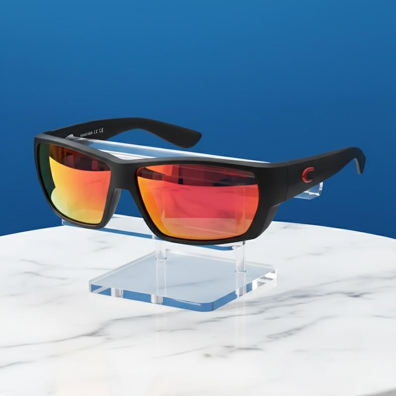 Puede incluir: Gafas de sol negras con lentes espejadas rojo-naranja se exhiben en un soporte de acr&iacute;lico transparente. Las gafas tienen una montura rectangular con un peque&ntilde;o logotipo rojo en la patilla. El fondo es un degradado de blanco y azul.