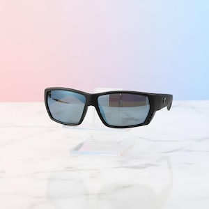 Puede incluir: Gafas de sol negras con lentes espejadas azules. Las gafas tienen una montura rectangular y se exhiben sobre un soporte de acr&iacute;lico transparente. El fondo presenta un degradado de rosa y azul.