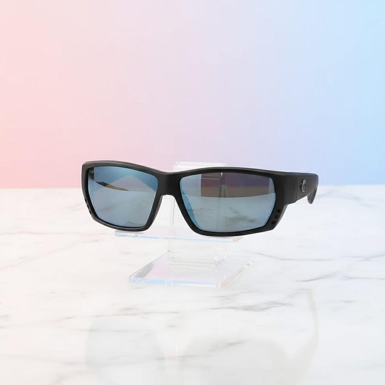 Puede incluir: Gafas de sol negras con lentes espejadas azules. Las gafas tienen una montura rectangular y se exhiben sobre un soporte de acr&iacute;lico transparente. El fondo presenta un degradado de rosa y azul.