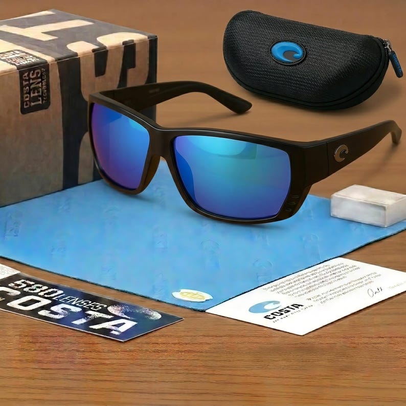 Puede incluir: Gafas de sol negras con lentes azules espejadas, un estuche negro y un pa&ntilde;o de limpieza. Las gafas tienen una montura rectangular y la marca "COSTA" es visible en el estuche y una tarjeta.
