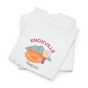 Puede incluir: Dos camisetas blancas dobladas con un diseño gráfico. El diseño presenta la palabra "Knoxville" en rosa, un estadio naranja, una bola colorida y la palabra "Tennessee" en verde.