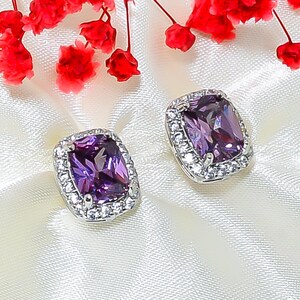 May include: A pair of silver-toned stud earrings with large, rectangular, purple gemstones. Each earring is surrounded by a halo of small, clear crystals. Red flowers are in the background, resting on a white, silky surface.