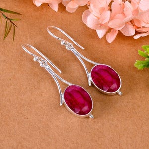 May include: Silver earrings featuring oval, faceted, ruby-red gemstones. The earrings have a hook design and a decorative silver frame. The earrings are displayed on a brown surface.