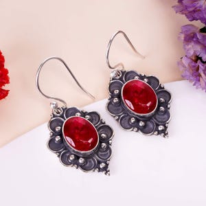 Peut inclure: Une paire de boucles d'oreilles en argent avec de grandes pierres précieuses ovales rouge foncé. Les pierres sont serties dans des montures argentées foncées ornées d'un motif floral. Les boucles ont une fermeture à crochet.