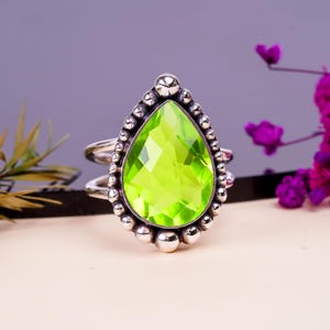 May include: A silver ring with a vibrant, pear-cut, lime green gemstone. The stone is framed by a beaded silver setting. The ring has a double band design. The background is a soft, neutral colour.