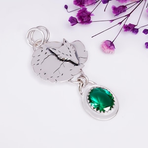 Puede incluir: Un colgante de plata que presenta un disco circular con un diseño de zorro grabado, conectado por una argolla a un engaste ovalado más pequeño que contiene una piedra preciosa de color verde esmeralda con talla brillante.