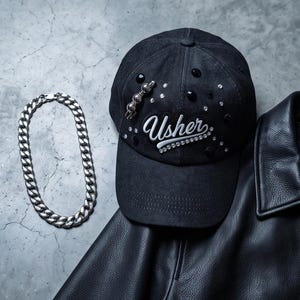 May include: A black baseball cap featuring the word Usher in white script embroidery, accented with scattered black and clear rhinestones and a silver-toned metal pin. Beside the cap is a thick silver-toned chain necklace and a black leather jacket.