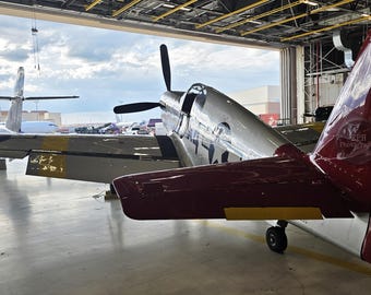 May include: Two vintage airplanes are parked inside a hangar. The closest plane has a silver body and a maroon tail with yellow accents. The other plane is olive green with a star emblem.