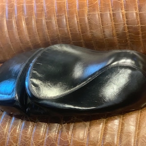 Peut inclure: Une selle de moto en cuir noir avec une finition brillante. La selle a une forme incurvée et est conçue pour s'adapter aux contours d'une moto. La selle est posée sur une surface en cuir marron.