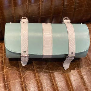 May include: A mint green cylindrical handbag with white straps and silver buckles. The bag features a white panel down the center and white straps with silver buckles. The bag is made of leather and has a rounded flap closure.
