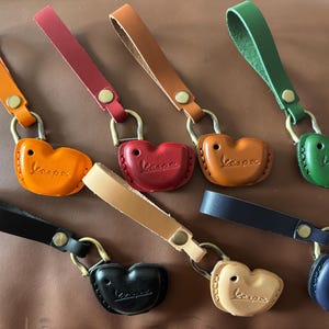 May include: A selection of leather keyrings in shades of orange, red, brown, green, black, beige, and navy. Each keyring has a heart-shaped design with the word "Vespa" embossed and a leather strap, attached with a metal clasp.