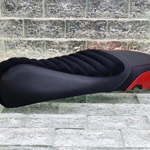 Może przedstawiać: Siedzenie motocyklowe z czarną skórzaną podstawą i czarnym, pionowo żebrowanym, przypominającym zamsz środkiem. Tylna część jest czerwona z czarnym akcentem. Widoczne są czerwone szwy. To niestandardowe siedzenie jest przeznaczone do motocykli.