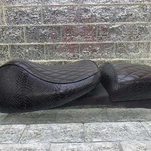 Może przedstawiać: Czarna siedzenie motocyklowe z pikowanym wzorem w romby i teksturowanym wzorem przypominającym skórę węża. Siedzenie to dwuczęściowy zestaw, składający się z większego siedzenia i mniejszego siedzenia pasażera. Siedzenie wykonane jest z trwałego materiału.