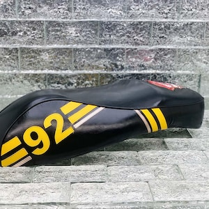 Może przedstawiać: Czarne siedzenie motocyklowe z żółtymi i białymi paskami oraz żółtym numerem "92". Siedzenie ma zakrzywiony kształt i czerwony emblemat na górze. Siedzenie jest wykonane z błyszczącego materiału.