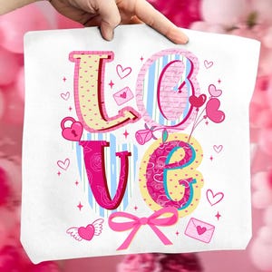 以下が含まれることがあります： 「LOVE」の文字がカラフルでロマンチックなデザインの白いトートバッグ。文字はハート、バラ、リボンで飾られています。「L」はピンク色で黄色の水玉模様、「O」は青いストライプです。