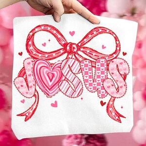 May include: White tote bag featuring the word "JESUS" in pink and red lettering, with a heart-shaped design for the letter "O". The design includes a large red bow and small hearts, perfect for Valentine's Day or religious gifts.