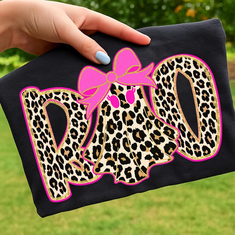 May include: Black t-shirt with a leopard print design spelling out the word "RAD". A pink bow and a leopard print ghost graphic are centered above the word. The design has gold glitter accents.