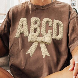 May include: Brown t-shirt with the letters ABCD in a cream-colored, knitted style, and a matching bow design. The shirt has a relaxed fit and a vintage aesthetic, perfect for casual wear.