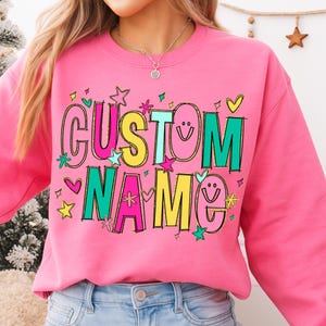 May include: A pink sweatshirt with the words "CUSTOM NAME" in colorful, outlined letters. The design includes stars, hearts, and smiley faces. The sweatshirt is a crew neck style.