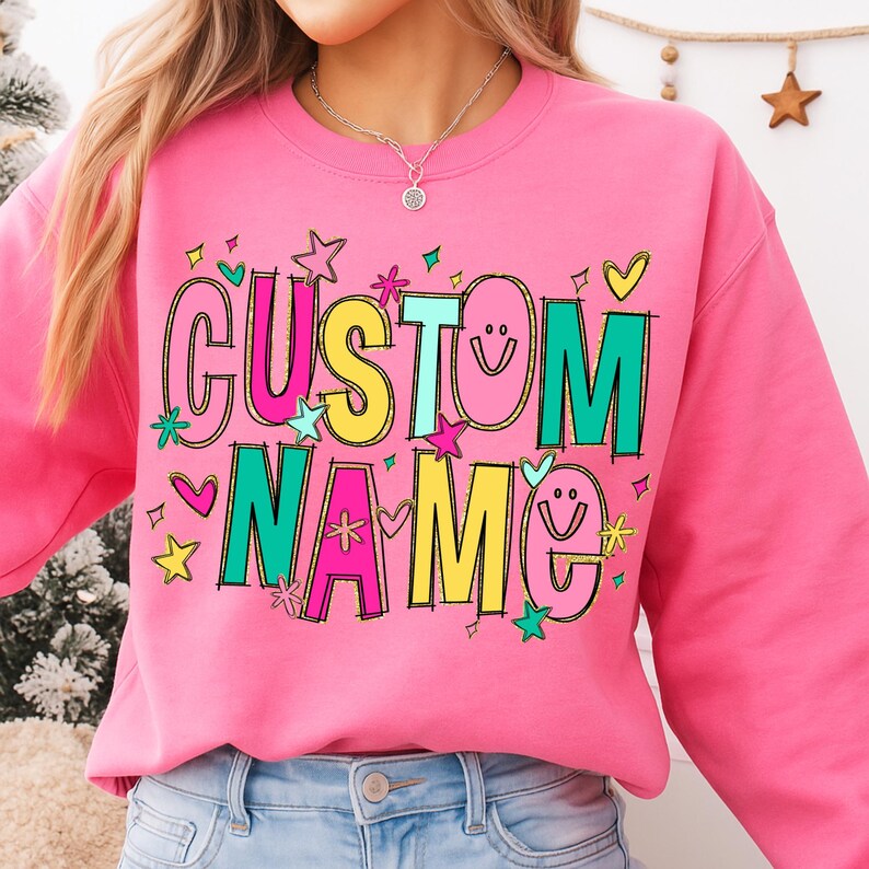 May include: A pink sweatshirt with the words "CUSTOM NAME" in colorful, outlined letters. The design includes stars, hearts, and smiley faces. The sweatshirt is a crew neck style.