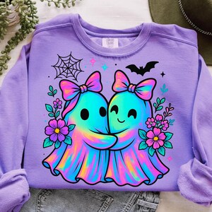 May include: A lilac-colored sweatshirt featuring a colorful graphic of two hugging ghosts with bows, surrounded by flowers, a spiderweb, and a bat. The ghosts are in shades of blue, pink, and yellow. The sweatshirt has long sleeves.