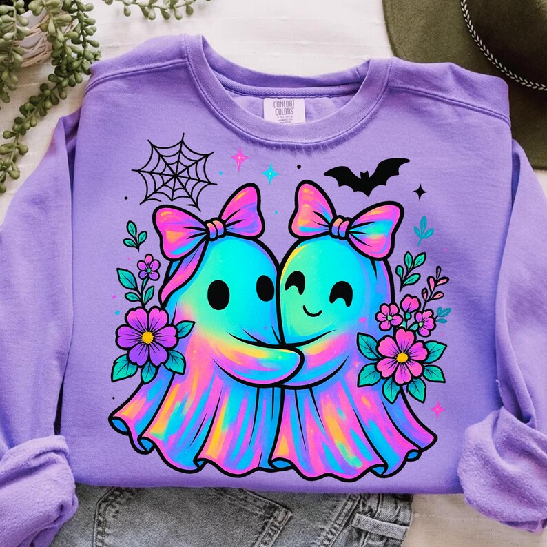 May include: A lilac-colored sweatshirt featuring a colorful graphic of two hugging ghosts with bows, surrounded by flowers, a spiderweb, and a bat. The ghosts are in shades of blue, pink, and yellow. The sweatshirt has long sleeves.