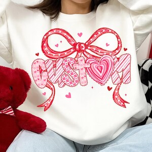 May include: White sweatshirt with a large red bow and the word "CUSTOM" in pink and red lettering. The design includes hearts and a candy cane stripe pattern, perfect for Valentine's Day or a custom gift.