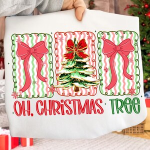 May include: White Christmas-themed sweatshirt with a festive design. It features three framed sections with a red bow, a Christmas tree, and the text "OH. CHRISTMAS TREE" in red and green. The design includes red, green, and pink accents.