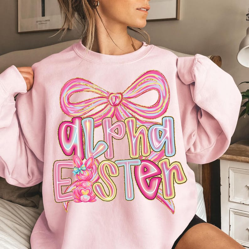 May include: A light pink sweatshirt with a large bow graphic and the words "Alpha Easter" in a colorful, outlined font. A cartoon bunny is also featured in the design. The sweatshirt is a long-sleeved, crew neck style.