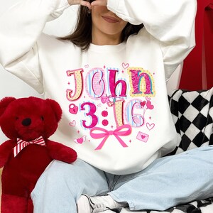 May include: White sweatshirt featuring the text "John 3:16" in a colourful, decorative font. The design includes hearts, a lock, and a pink bow. A red teddy bear is visible on the left.