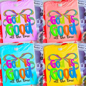 May include: Four colorful t-shirts: pink, coral, blue, and yellow. Each shirt features the text "God is Good all the time" in vibrant, multi-colored letters and a decorative bow design. The shirts are displayed on a patterned surface.