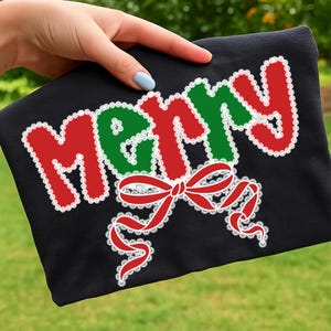 May include: Black t-shirt with the word "MERRY" in red and green, outlined with white lace. A red bow with white lace accents is below the word. The shirt is held by a hand with a light blue nail polish.