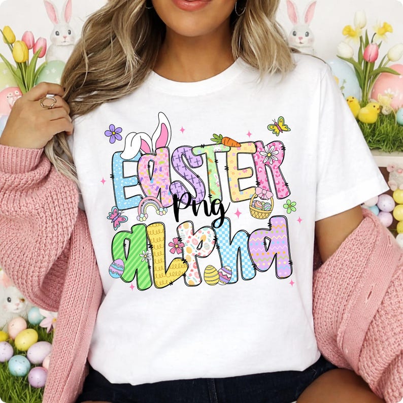 Op de afbeelding: Wit T-shirt met de tekst "Easter Png Alpha" in kleurrijke, patroonletters. Het ontwerp bevat paaseieren, vlinders, bloemen en een regenboog. Een feestelijk ontwerp met een lentethema.